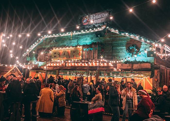 A crowded Christmas market called “Bavarian Bar” at night, surrounded by lights and people drinking